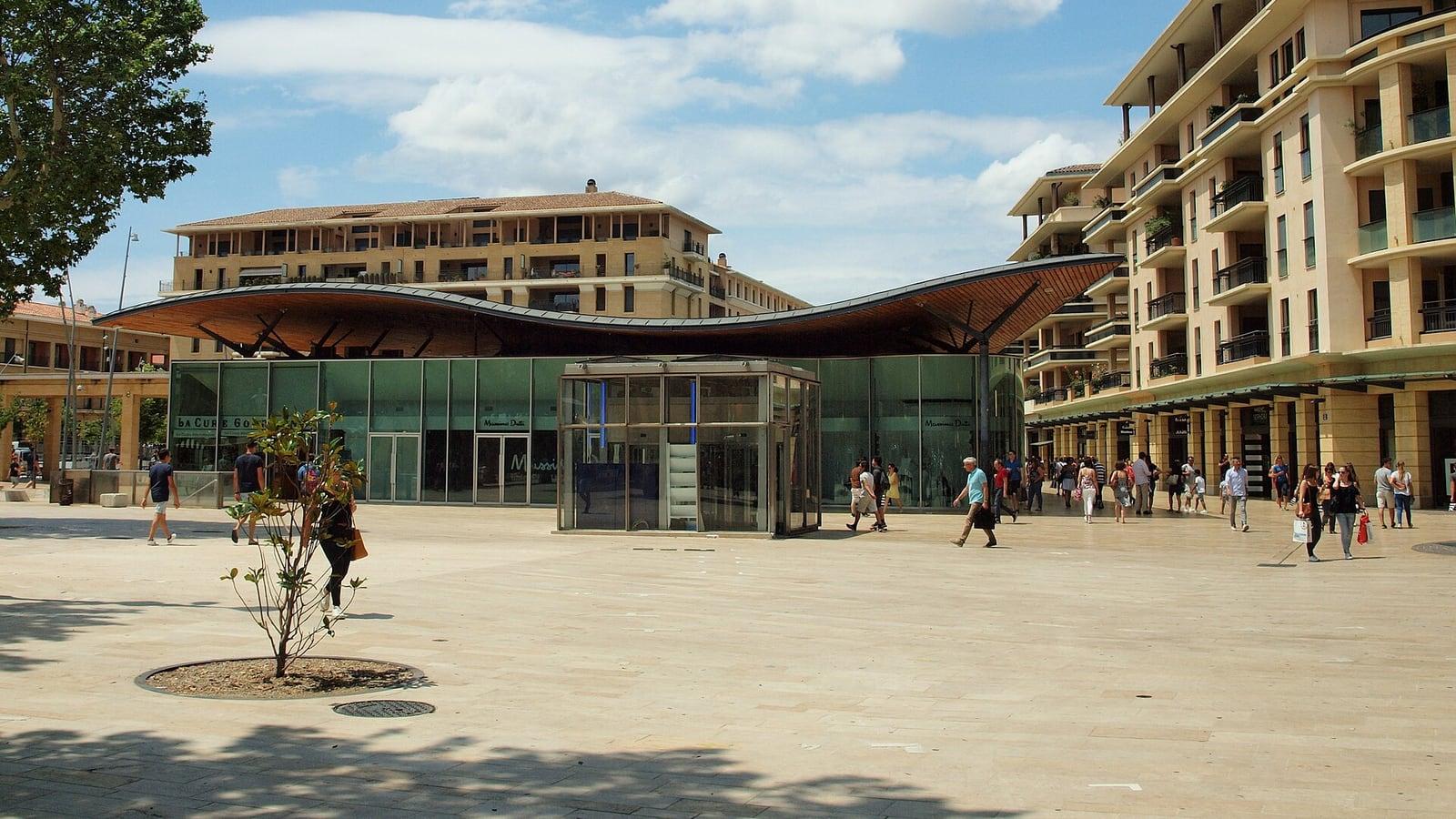 L'esplanade piétonne des Allées Provençales à Aix-en-Provence, son pavillon vitré sous une toiture ondulée et des immeubles crème, avec quelques passants