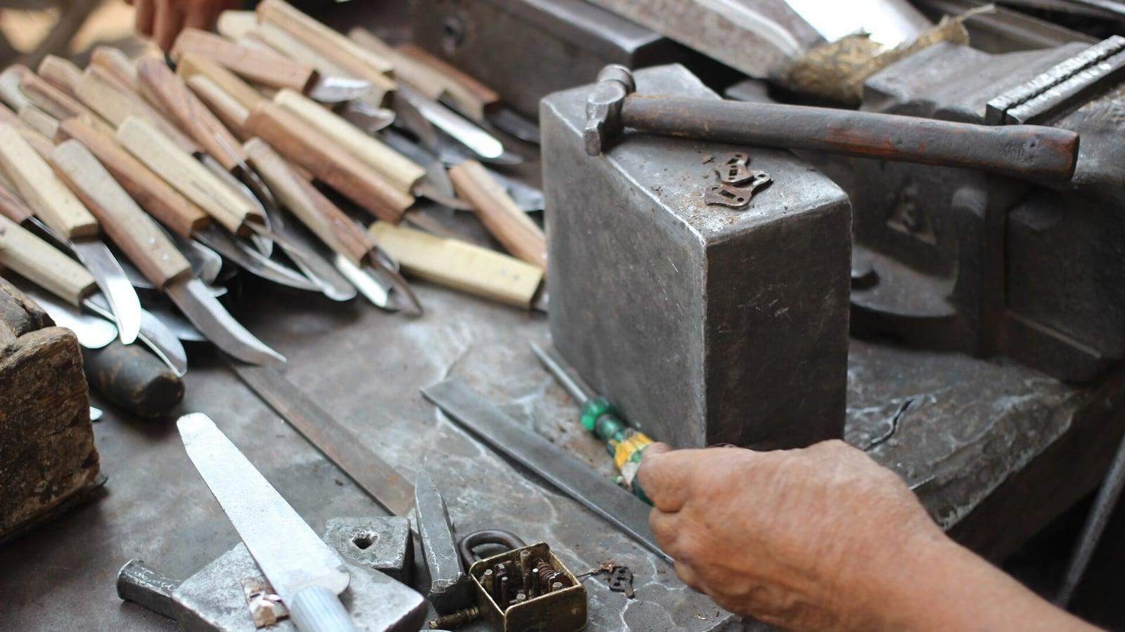 Établi d'un coutelier : lames à manche de bois, enclume, marteau et outils, une main tenant un tournevis