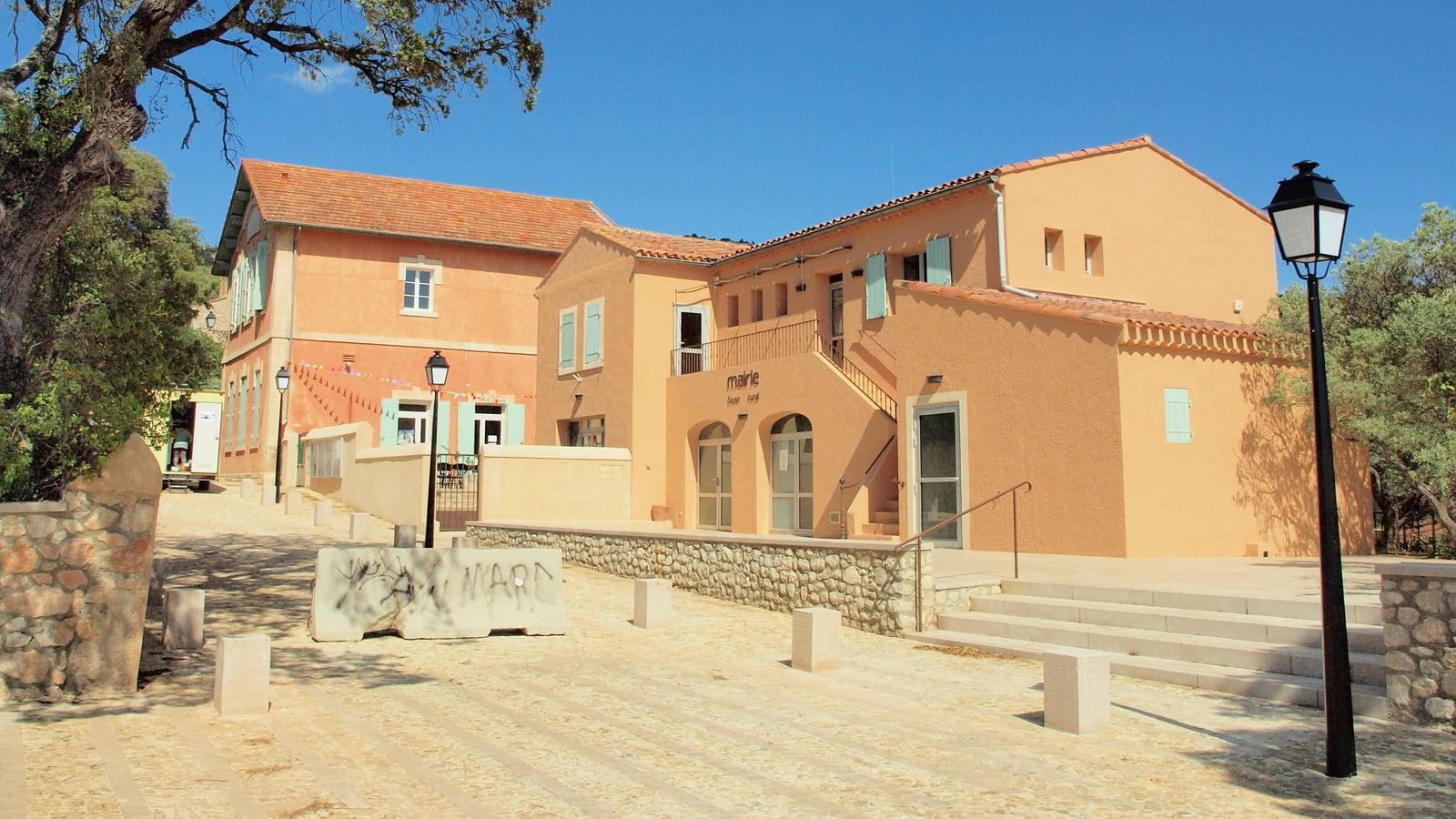 Le bâtiment de la mairie et de l'école de Beaurecueil, aux façades ocre et volets vert d'eau, devant une place pavée bordée de réverbères