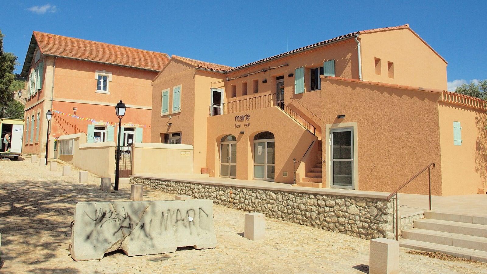 Le bâtiment ocre de la mairie et de l'école de Beaurecueil, avec son escalier extérieur, vu depuis la place sous un ciel bleu