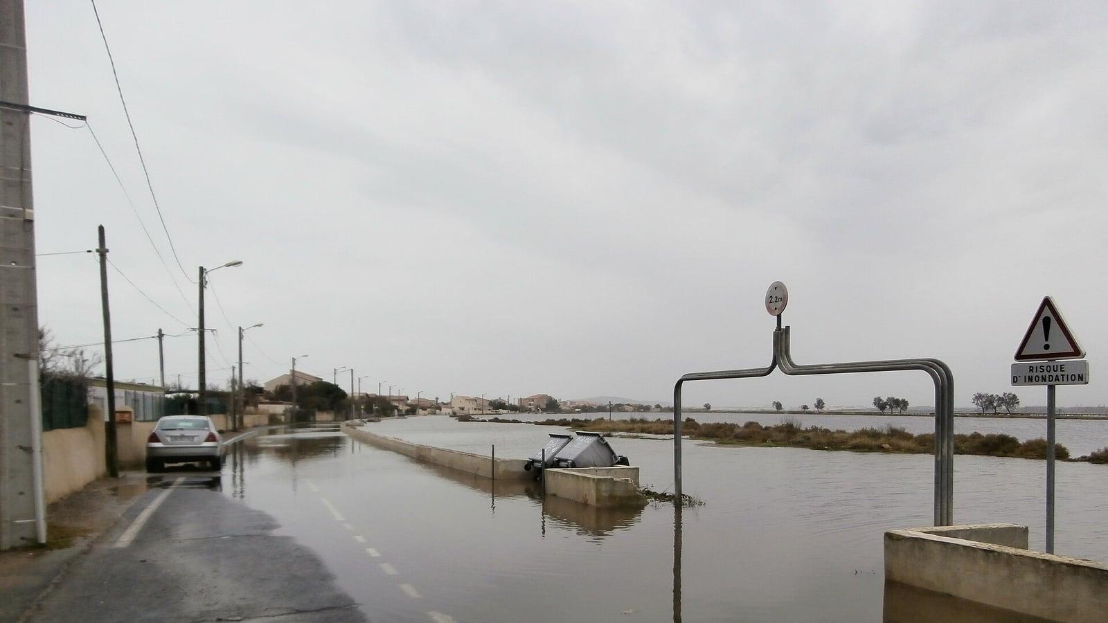 Une route inondée sous un ciel gris, une voiture garée au bord de l'eau et un panneau « Risque d'inondation » à droite