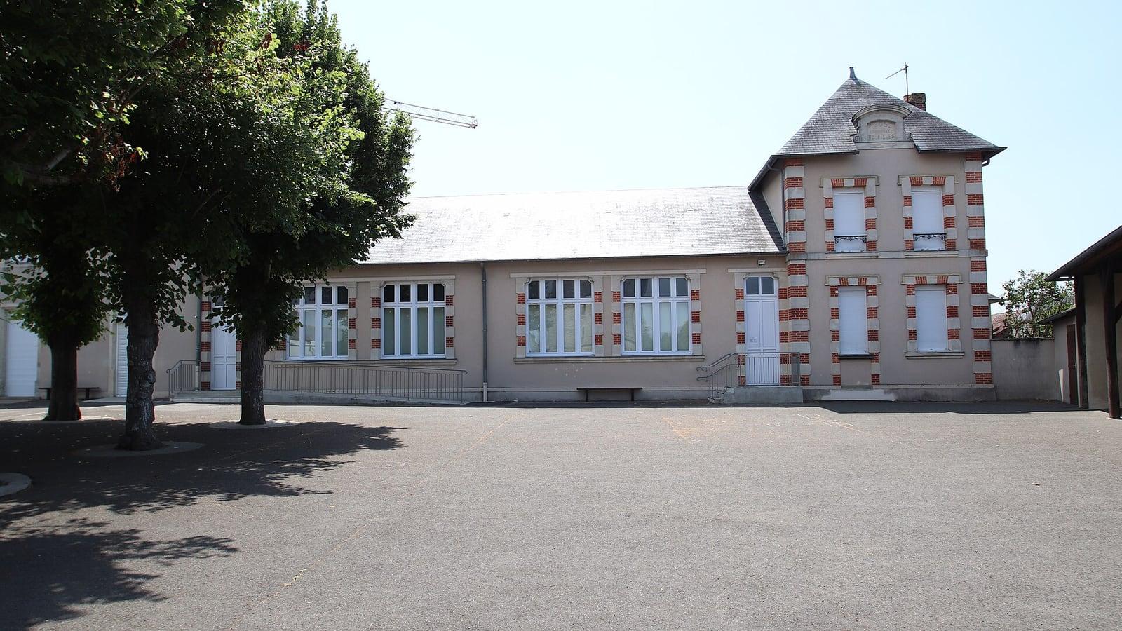 La cour goudronnée d'une école primaire en plein soleil, ombragée en partie par trois arbres, devant un bâtiment scolaire en brique et pierre (école de Cherré-Au, dans la Sarthe)