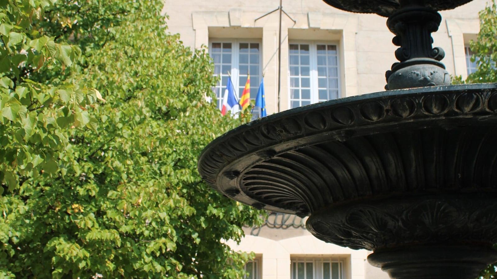 Façade de la mairie de Cabriès, ses drapeaux français, provençal et européen au balcon, derrière un arbre et une fontaine en fonte