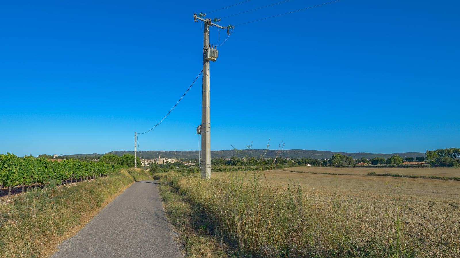Un chemin goudronné entre une vigne et un champ moissonné, longé par un poteau électrique en béton qui porte un petit transformateur, sous un ciel bleu