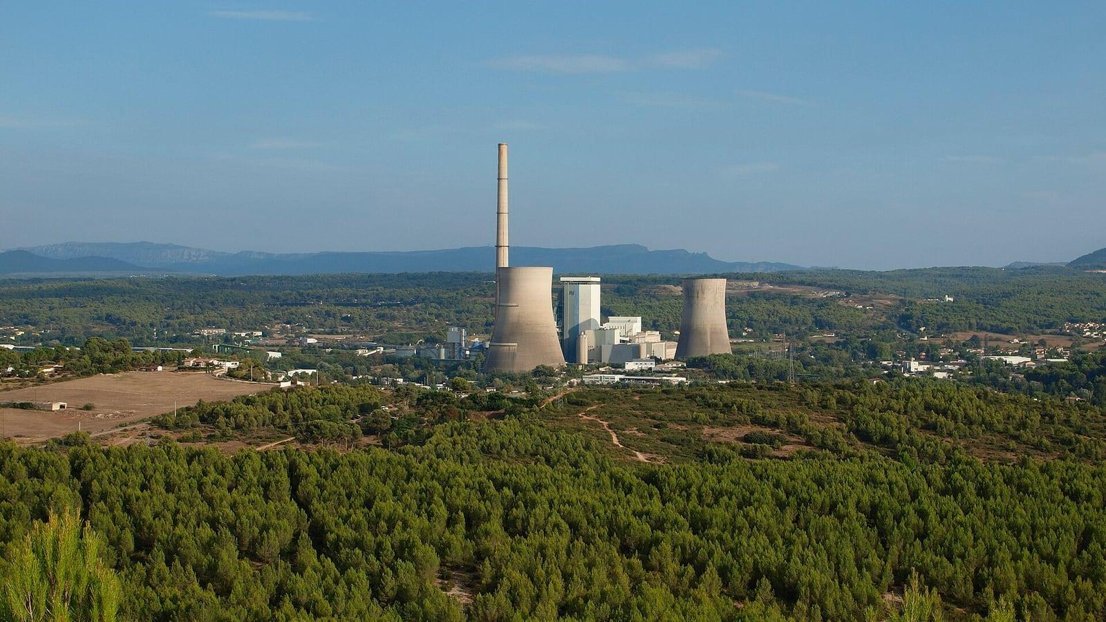 La centrale de Provence, sa haute cheminée et ses deux tours de refroidissement, vue au-dessus d'une pinède, avec des reliefs à l'horizon
