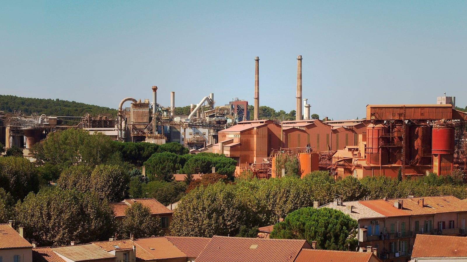 Les bâtiments rouillés, les silos et les deux hautes cheminées de l'usine d'alumine de Gardanne, derrière une rangée d'arbres et des toits de tuiles