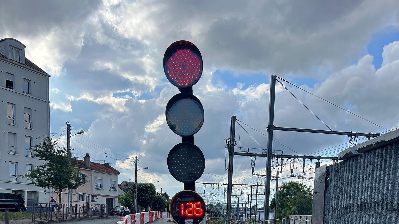 Un feu tricolore de chantier au rouge, avec un compte à rebours lumineux, devant des barrières rouges et blanches au bord d'une rue