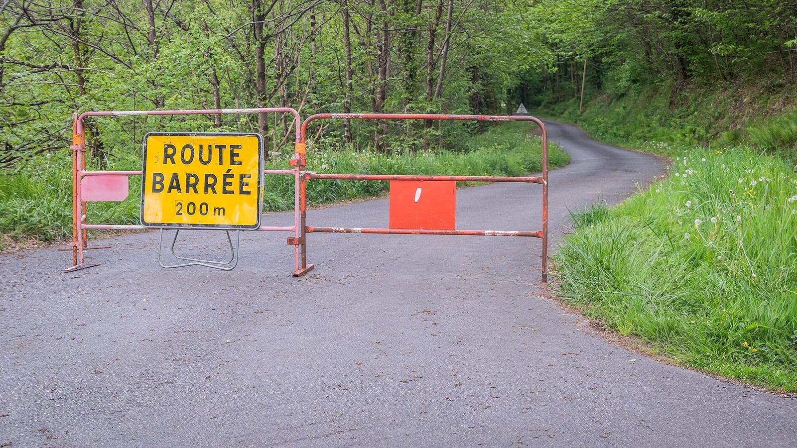 Une petite route bordée d'arbres, fermée par des barrières de chantier rouges et un panneau jaune « Route barrée 200 m »