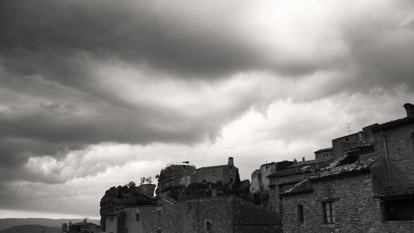 Un ciel d'orage chargé de nuages sombres au-dessus des maisons en pierre d'un village perché de Provence, en noir et blanc