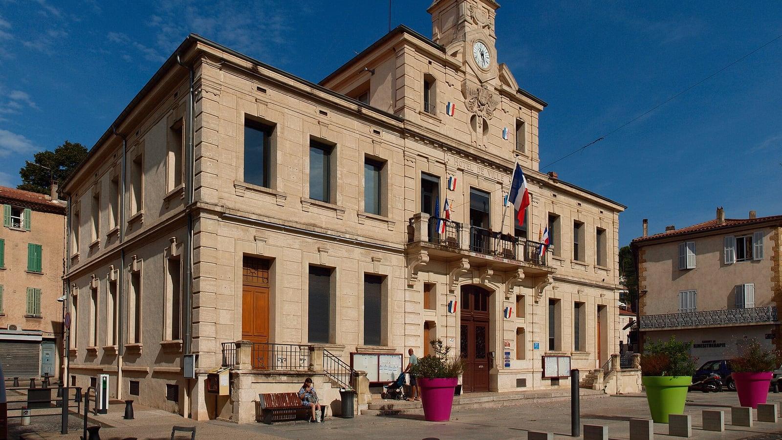 La façade en pierre de l'hôtel de ville de Gardanne, avec son horloge et ses drapeaux, sous un ciel bleu