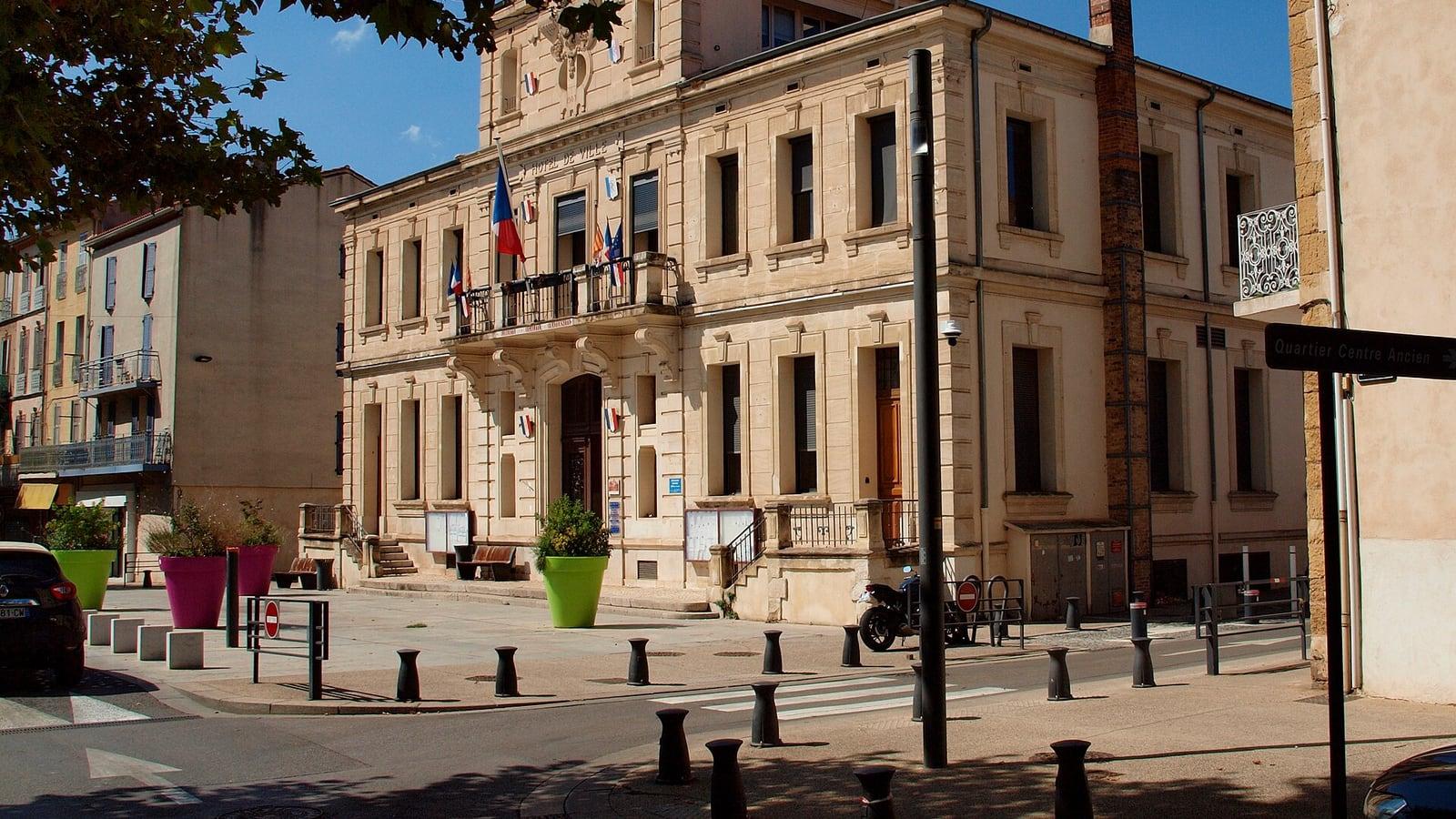 La façade en pierre claire de l'hôtel de ville de Gardanne, avec ses drapeaux au balcon, vue depuis un carrefour bordé de potelets