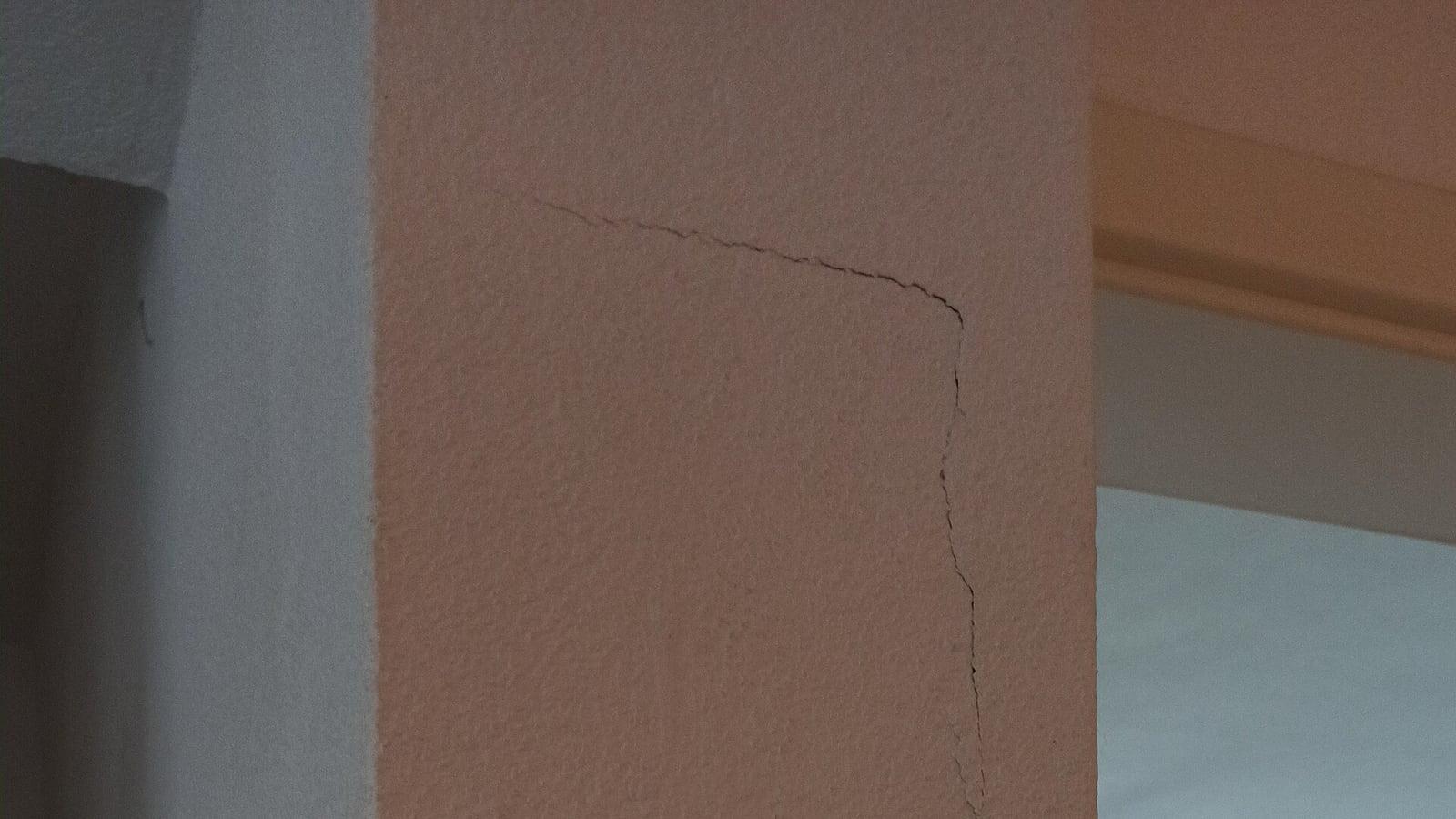 Une fissure fine court en angle sur un mur intérieur crépi de couleur saumon, près de l'encadrement d'une porte