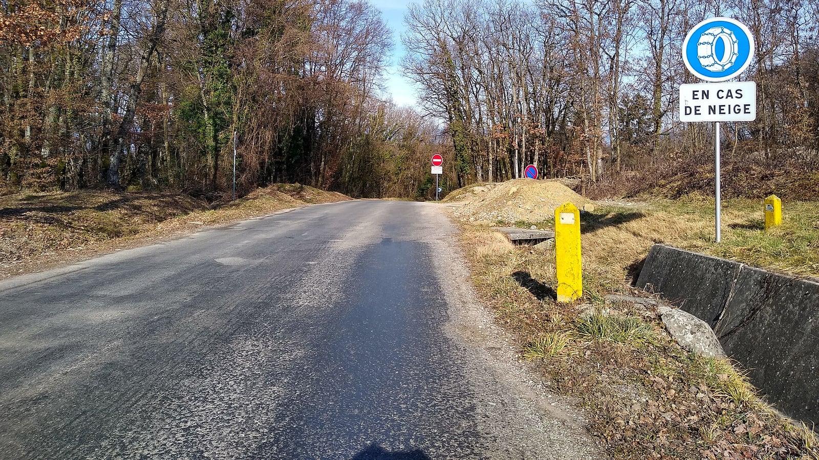 Une petite route goudronnée entre des arbres sans feuilles, avec au bord un panneau rond bleu figurant une roue équipée de chaînes et le panonceau « En cas de neige »