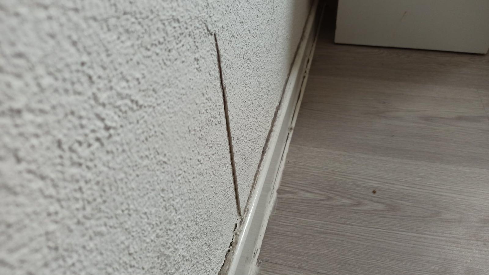 Une fissure fine court en diagonale sur un mur crépi blanc, jusqu'à la plinthe, au-dessus d'un parquet clair