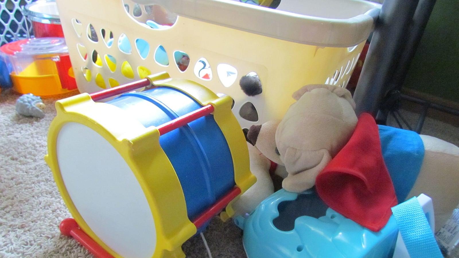 Jouets de tout-petit posés sur une moquette : un tambour jaune et bleu, une peluche de chien, un jouet en plastique bleu et un panier à linge ajouré