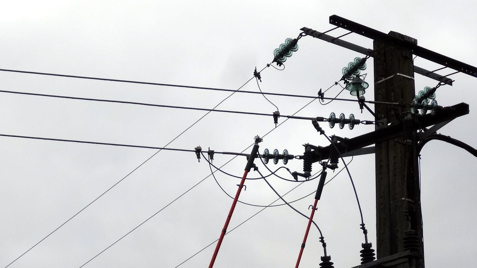 Le haut d'un poteau électrique avec ses isolateurs et ses câbles, deux perches rouges d'intervention tendues vers la ligne, sous un ciel gris