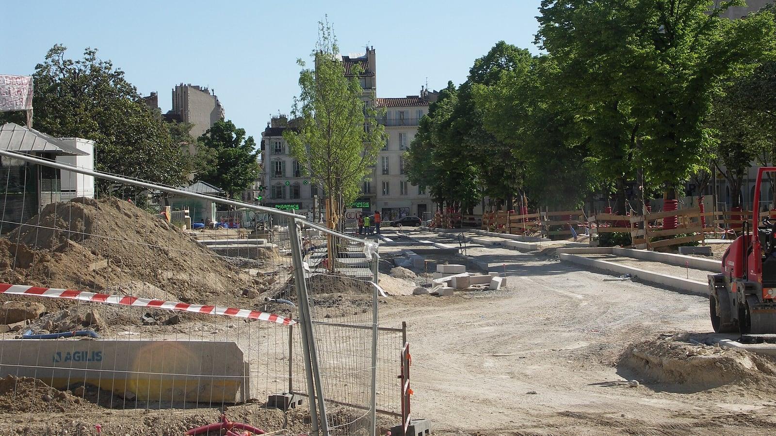 Chantier de voirie derrière des barrières métalliques et un ruban rouge et blanc à Marseille, avec des tas de terre, un engin de travaux et quelques ouvriers en tenue orange au loin