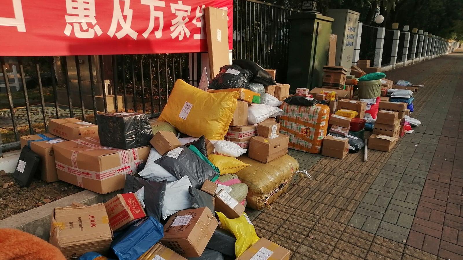 Des dizaines de colis en carton et de sachets plastiques entassés sur un trottoir le long d'une grille, sous une banderole rouge écrite en chinois