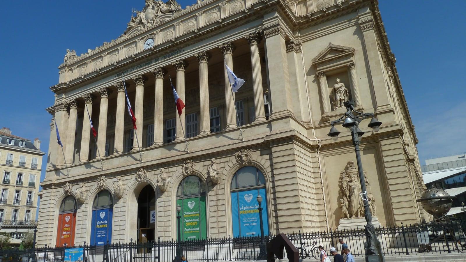 Façade à colonnes du palais de la Bourse, siège de la chambre de commerce à Marseille, avec des drapeaux et des bannières colorées sous les arcades, sous un ciel bleu