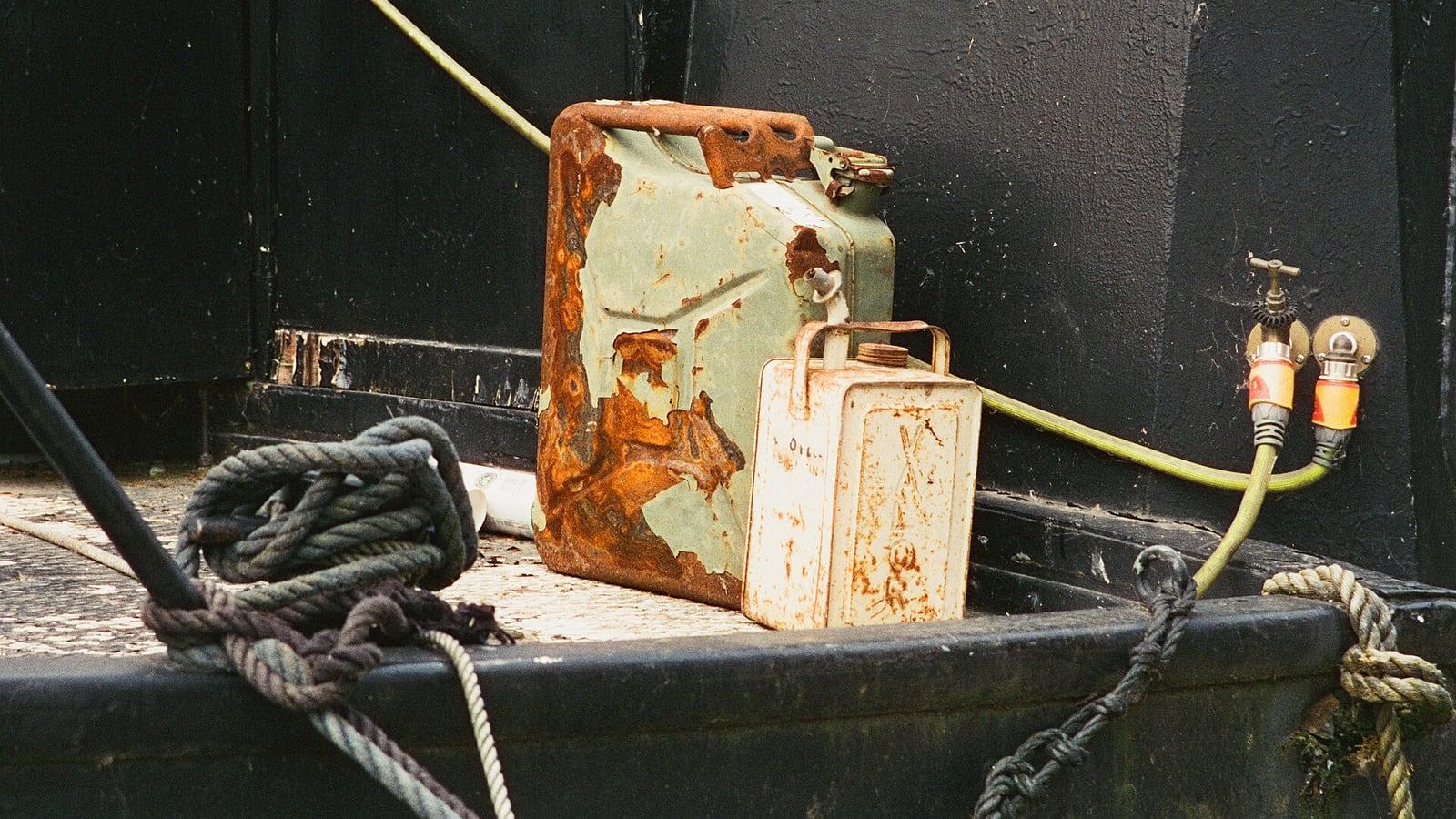 Un vieux jerrican métallique vert piqué de rouille et un petit bidon blanc posés sur le pont d'un bateau, entre des cordages