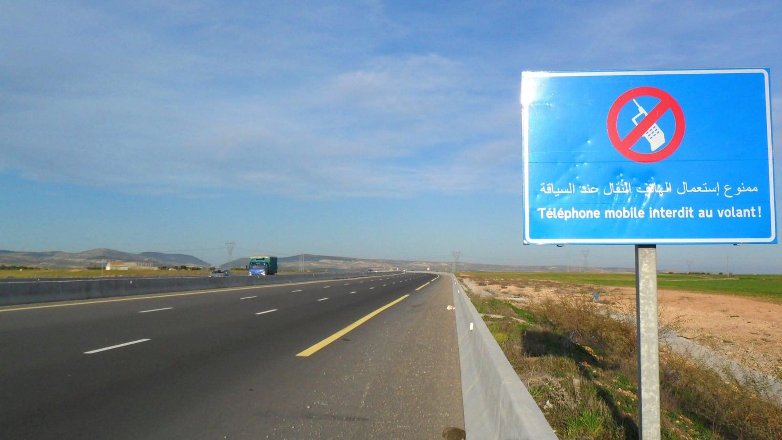 Panneau bleu au bord d'une autoroute à deux fois deux voies, barré d'un téléphone et portant l'inscription « Téléphone mobile interdit au volant ! » en arabe et en français
