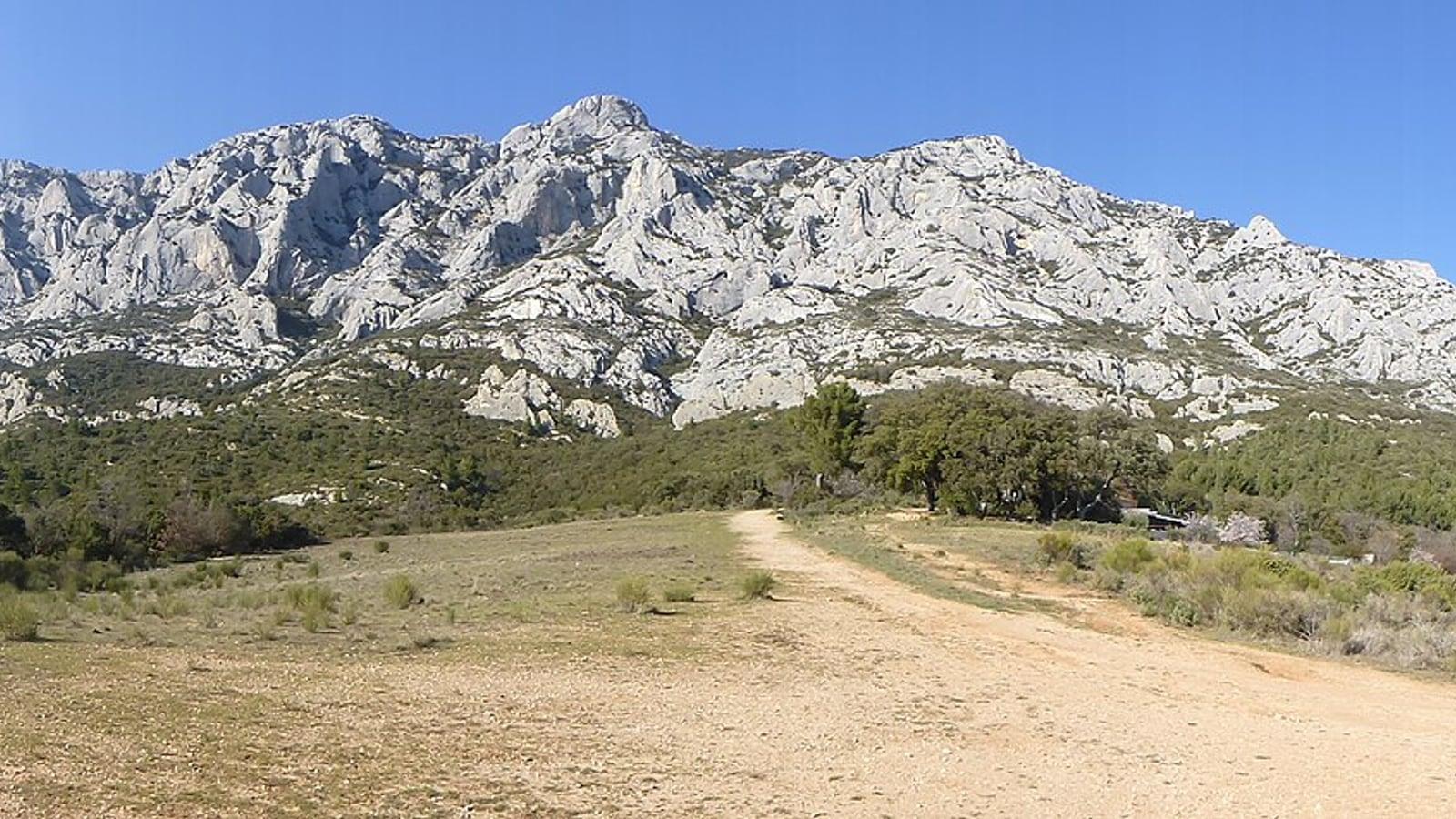 Une piste de terre traverse une clairière au pied des falaises calcaires de la face sud de Sainte-Victoire, sous un ciel bleu