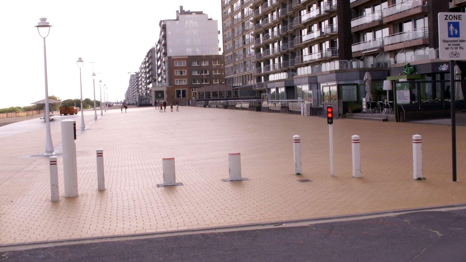 Bornes blanches à bandes rouges et feu rouge à l'entrée d'une esplanade piétonne pavée, devant des immeubles en bord de mer, à Coxyde en Belgique