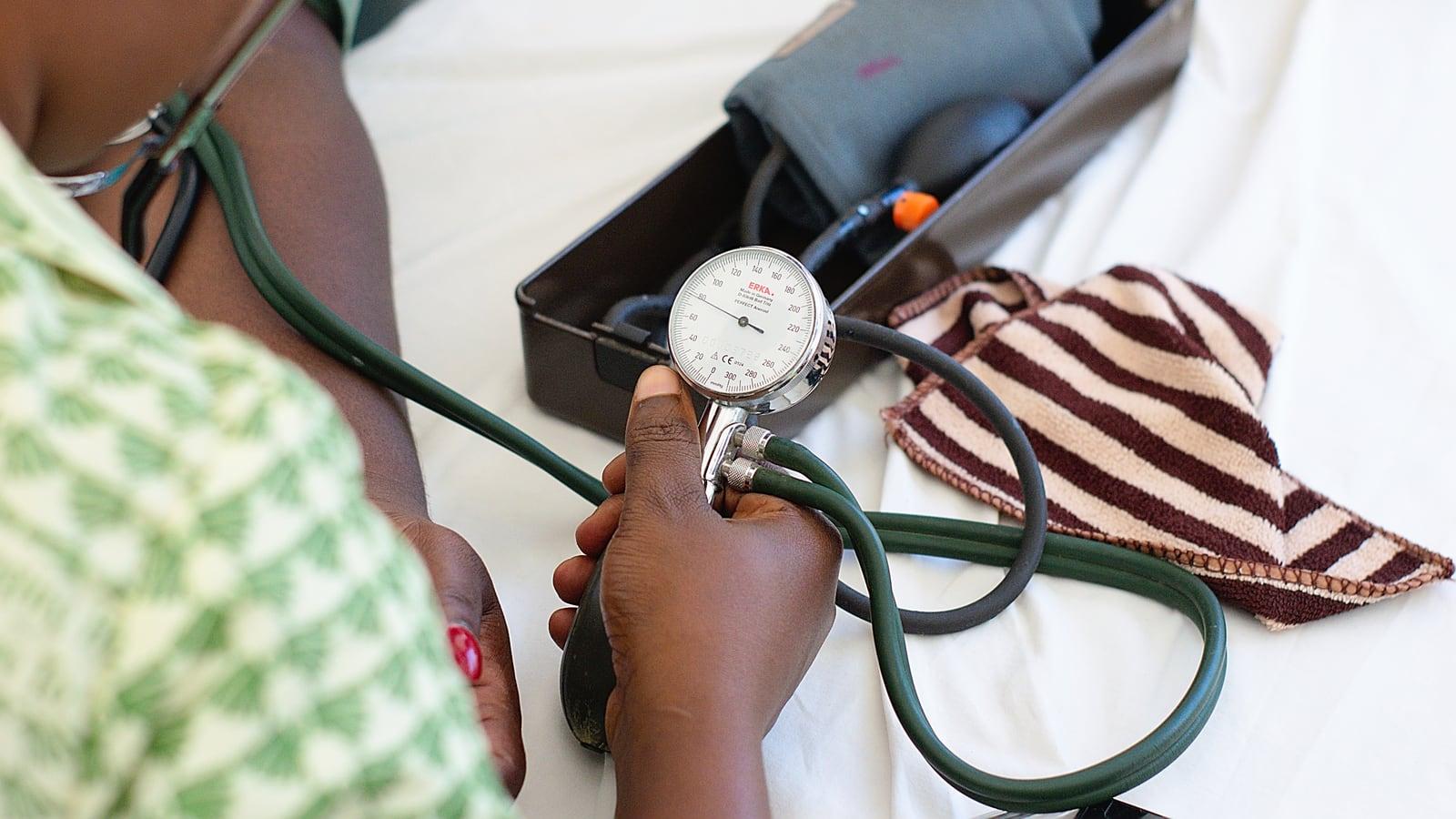 Une personne en blouse à motifs verts tient le manomètre d'un tensiomètre près du bras d'un patient, au-dessus d'un drap blanc