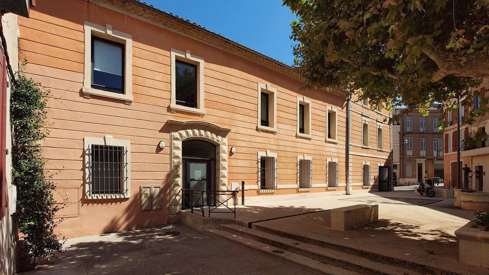 Façade ocre d'un bâtiment de la mairie de Trets aux fenêtres encadrées de pierre, sur une placette ombragée par un platane